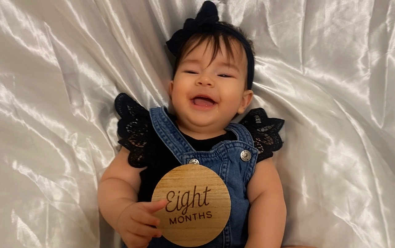Aurora joined the competition — help win amazing prizes! baby, infant, smiling, denim_overalls, black_clothing, lace, headband, bow, wooden_sign, eight_months, hand, lying_down, white_satin_background, cute, happy, portrait, child, skin, face, person