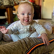 Layton joined the competition — help win amazing prizes! baby, child, infant, portrait, sitting, smiling, tongue_out, blue_eyes, blond_hair, onesie, indoors, home, cushion, pillow, bolster, bed, furniture, playful, cozy, closeup