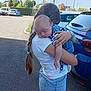asphalt, baby, blue_jeans, braid, car, casual_clothing, child, daylight, girl, hand, holding, nature, outdoor, pacifier, parking_lot, person, sleeping, sunny, tree, white_tshirt