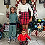 bicycle, celebration, child, children, christmas_tree, decorations, family, festive, green_outfit, happy, holiday, indoor, presents, red_skirt, sitting, smile, standing, tiled_floor, toys, wall_art