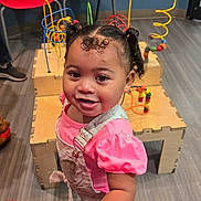 Patience joined the competition — help win amazing prizes! bead_maze, child, colorful_chairs, curly_hair, cute, flooring, happy, indoor, overalls, person, pigtails, pink_shirt, playroom, reaching, smiling, standing, toddler, toys, wooden_toy, young_child