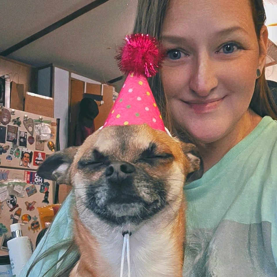 Olive is registered to the contest to win money with this photo: woman, dog, party_hat, indoor, smiling, pet, celebration, kitchen, refrigerator, magnets, casual_clothing, short_hair, light_skin, facial_expression, portrait, happy, animal, headshot, home, human