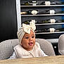 Manël participe au concours pour gagner de l'argent avec cette photo : baby, child, headwrap, bow, vest, high_chair, table, wine_bottles, shelf, indoor, smiling, happy, face, person, clothing, furniture, portrait, cute, seated, curious