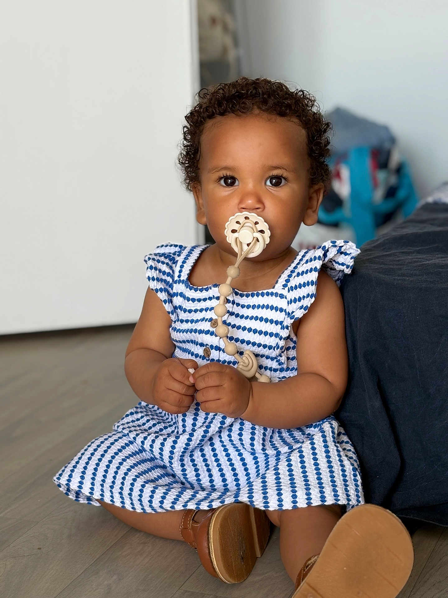Manël participe au concours pour gagner de l'argent avec cette photo : toddler, child, pacifier, dress, blue_and_white, curly_hair, sitting, floor, indoor, shoes, brown_skin, cute, portrait, baby, clothing, person, young_child, indoors, footwear, looking_at_camera