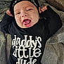 baby, infant, smiling, black_clothing, hat, text_on_clothing, blanket, lying_down, happy, cute, tongue_out, indoors, cozy, face, child, young, person, portrait, headwear, soft_texture