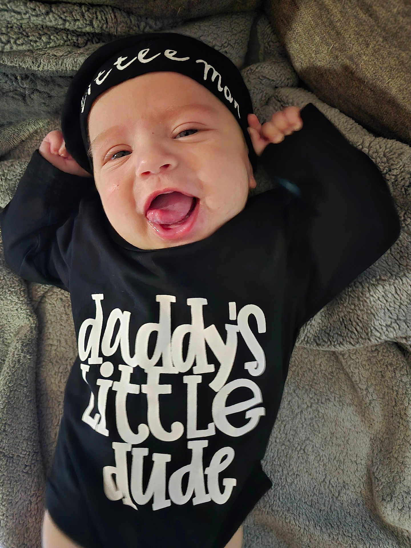 Hudson joined the competition — help win amazing prizes! baby, infant, smiling, black_clothing, hat, text_on_clothing, blanket, lying_down, happy, cute, tongue_out, indoors, cozy, face, child, young, person, portrait, headwear, soft_texture