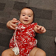 Miracle is registered to the contest to win money with this photo: baby, child, infant, red_clothing, floral_pattern, pacifier, carpet, reaching_hands, smiling, cute, candid, lying_down, happy, indoors, person, young_child, skin, face, toddler, playful
