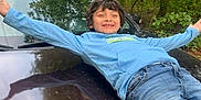 Bryson is registered to the contest to win money with this photo: child, boy, car, hood, jeans, blue_shirt, outdoor, trees, forest, smiling, happy, arms_outstretched, reflection, nature, casual_clothing, daylight, leaves, person, vehicle, relaxed