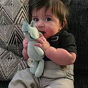 Cristian joined the competition — help win amazing prizes! baby, child, infant, plush_toy, stuffed_animal, couch, pillow, geometric_pattern, clothing, pants, shirt, barefoot, indoor, curly_hair, cute, sitting, face, hand, toys, home