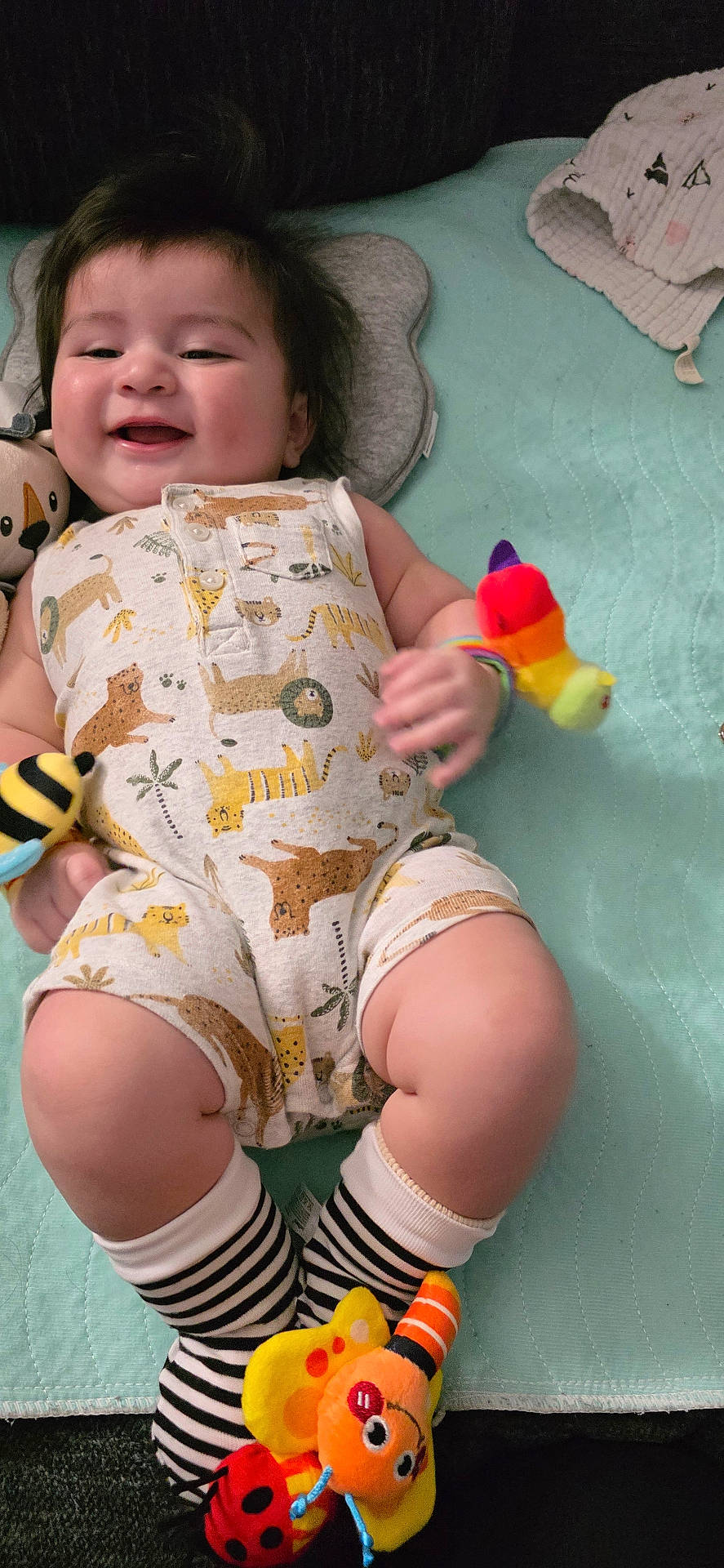 animal_print, baby, child, colorful, comfort, cute, face, happy, indoors, infant, lying_down, person, playful, plush_toy, romper, small_hands, smiling, soft_blanket, striped_socks, toy