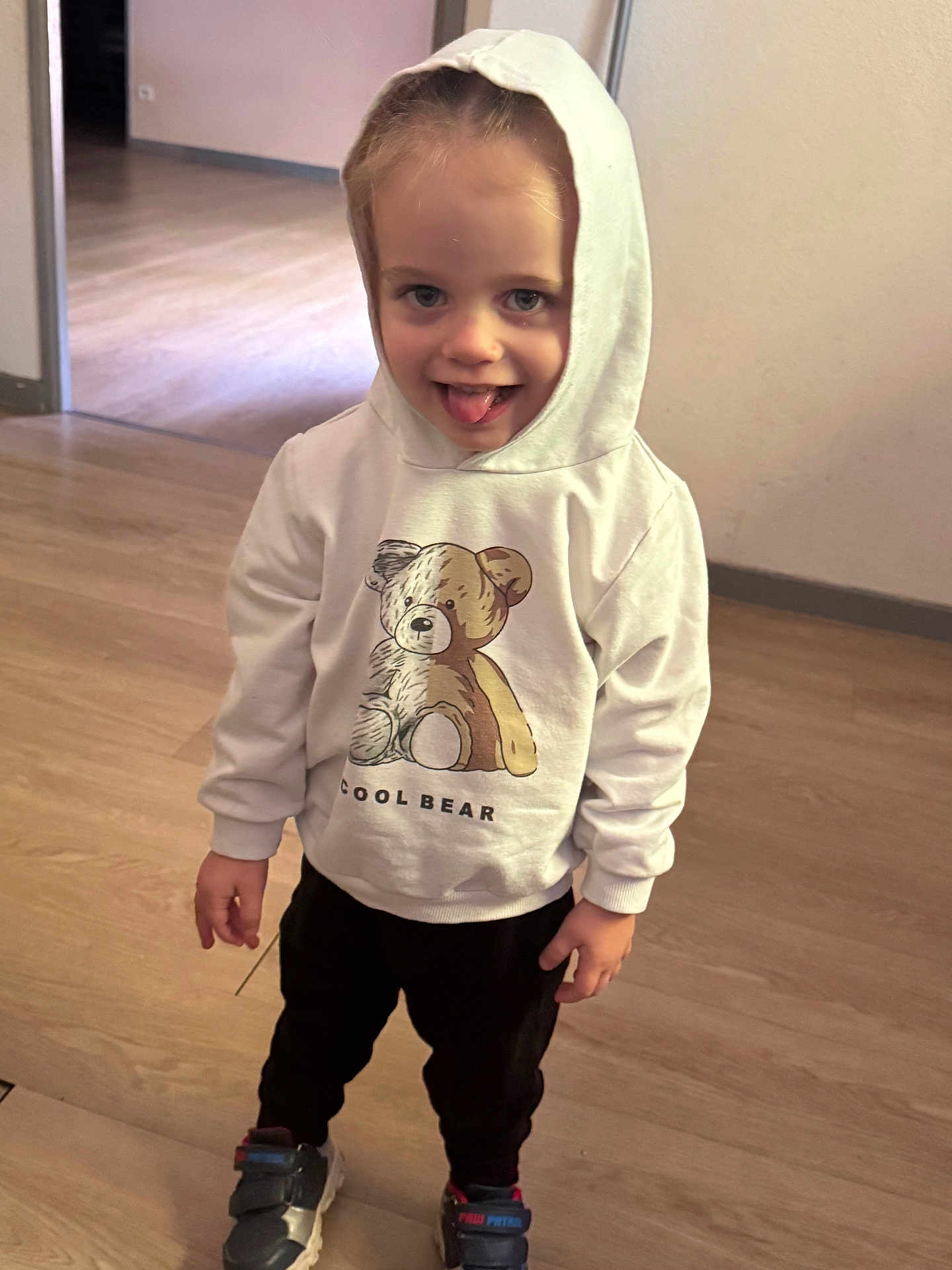 Melvyn a rejoint le concours — aidez-le/la à gagner de superbes lots ! toddler, child, hoodie, white_hoodie, teddy_bear, playful, tongue_out, indoor, wooden_floor, black_pants, sneakers, person, smiling, cute, young_child, casual_clothing, standing, light_skin, short_hair, happy