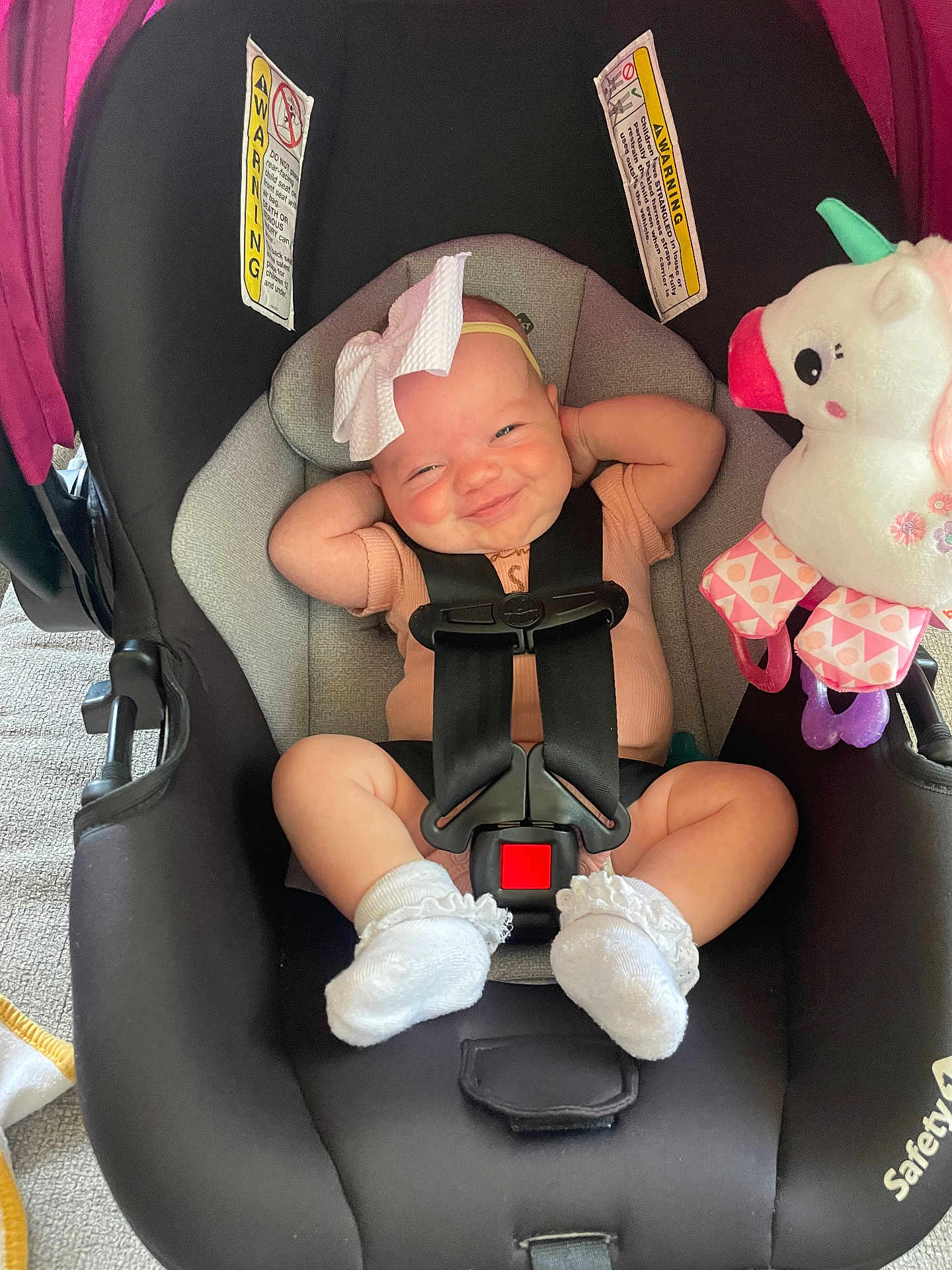 Laylah is registered to the contest to win money with this photo: baby, car_seat, smiling, bow, infant, toy, unicorn, plush, socks, straps, seatbelt, cute, child, indoor, car_seat_warning, pink, black, gray, comfort, happy