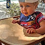 toddler, child, high_chair, cap, shirt, wooden_furniture, outdoor, daylight, tree, leafy, curious, blue_eyes, shorts, striped_shorts, hand, face, portrait, sunlight, summer, nature