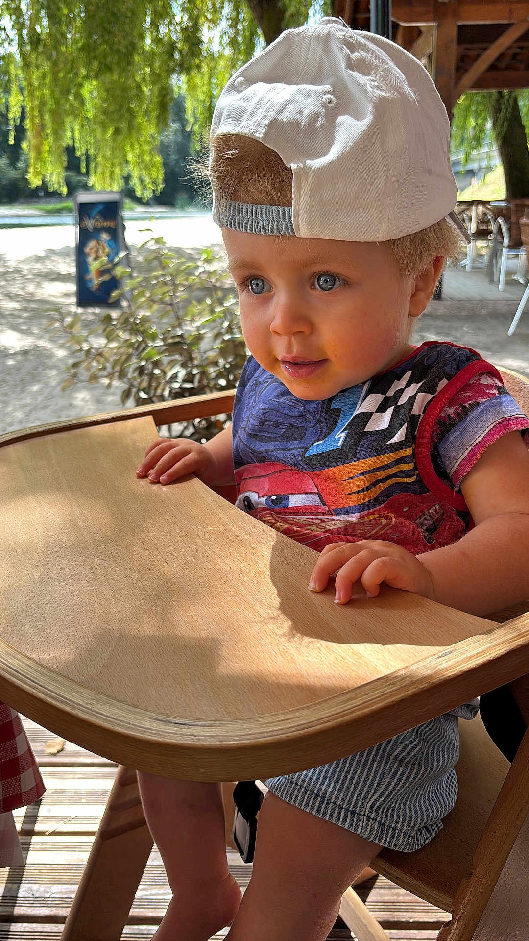 Sacha participe au concours pour gagner de l'argent avec cette photo : toddler, child, high_chair, cap, shirt, wooden_furniture, outdoor, daylight, tree, leafy, curious, blue_eyes, shorts, striped_shorts, hand, face, portrait, sunlight, summer, nature