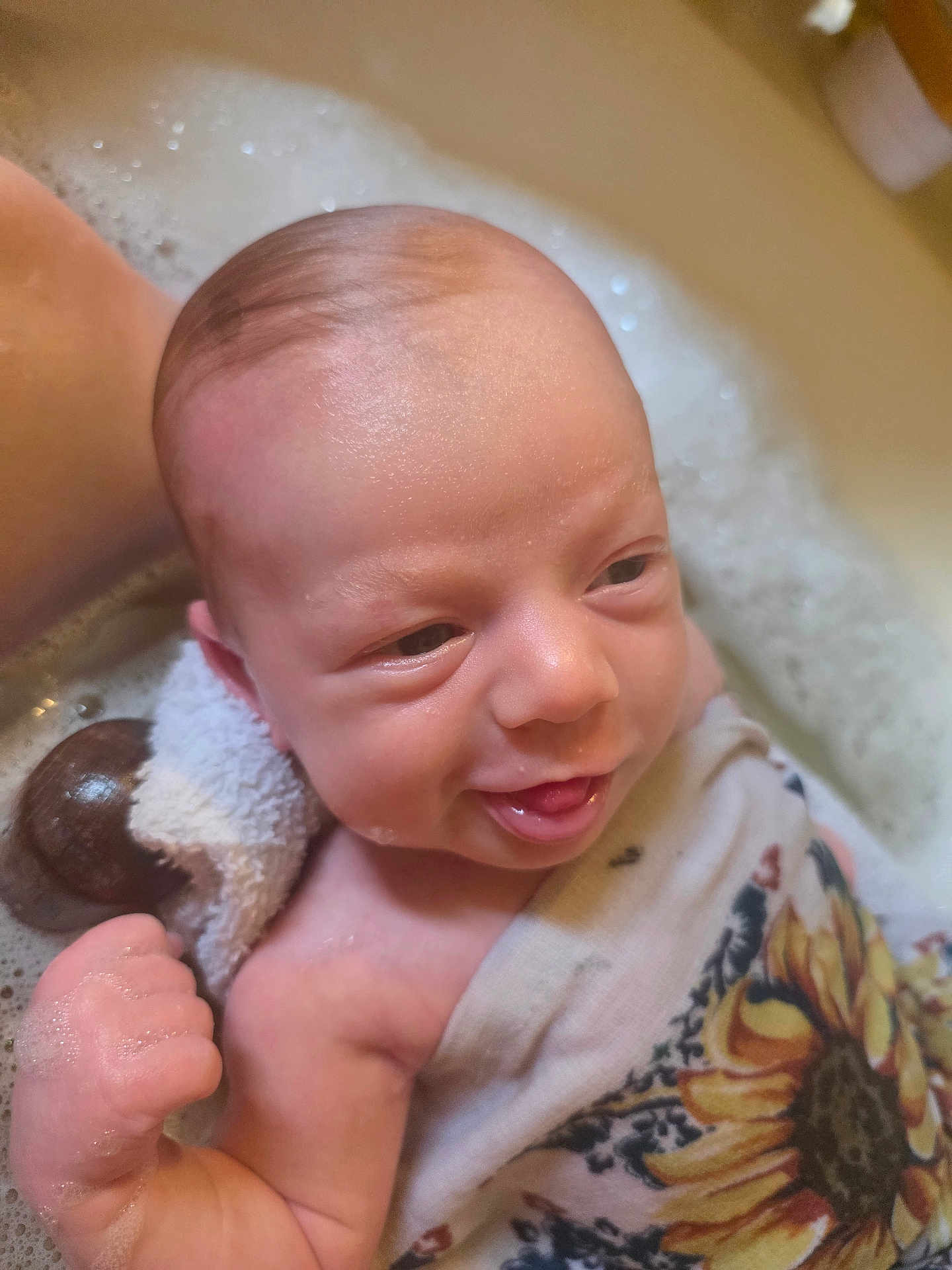Nova is registered to the contest to win money with this photo: baby, infant, newborn, bathtub, bubbles, towel, foam, water, bath_time, face, head, hand, smile, tongue, skin, closeup, cloth_pattern, sunflower_pattern, happy, relaxed
