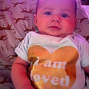 Aurora is registered to the contest to win money with this photo: baby, infant, child, face, blue_eyes, white_shirt, golden_heart, text_on_clothing, pillow, leopard_print, cute, chubby_cheeks, lying_down, indoor, soft_lighting, skin, person, portrait, toddler, love_message