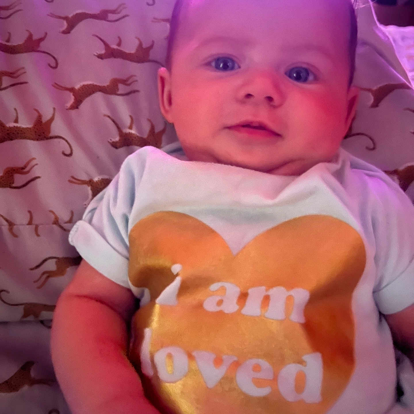 Aurora is registered to the contest to win money with this photo: baby, blue_eyes, child, chubby_cheeks, cute, face, golden_heart, indoor, infant, leopard_print, love_message, lying_down, person, pillow, portrait, skin, soft_lighting, text_on_clothing, toddler, white_shirt