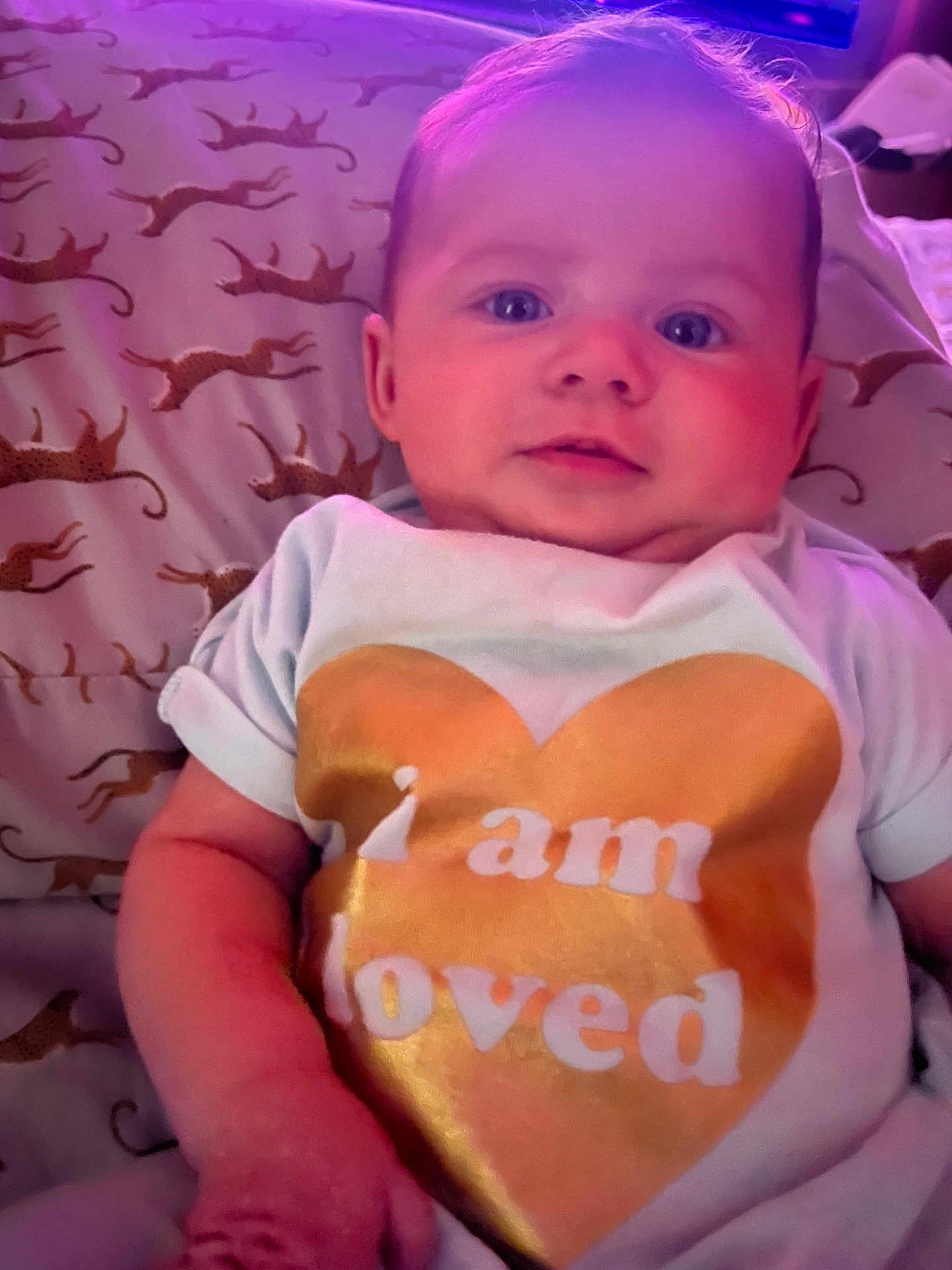 Aurora is registered to the contest to win money with this photo: baby, infant, child, face, blue_eyes, white_shirt, golden_heart, text_on_clothing, pillow, leopard_print, cute, chubby_cheeks, lying_down, indoor, soft_lighting, skin, person, portrait, toddler, love_message