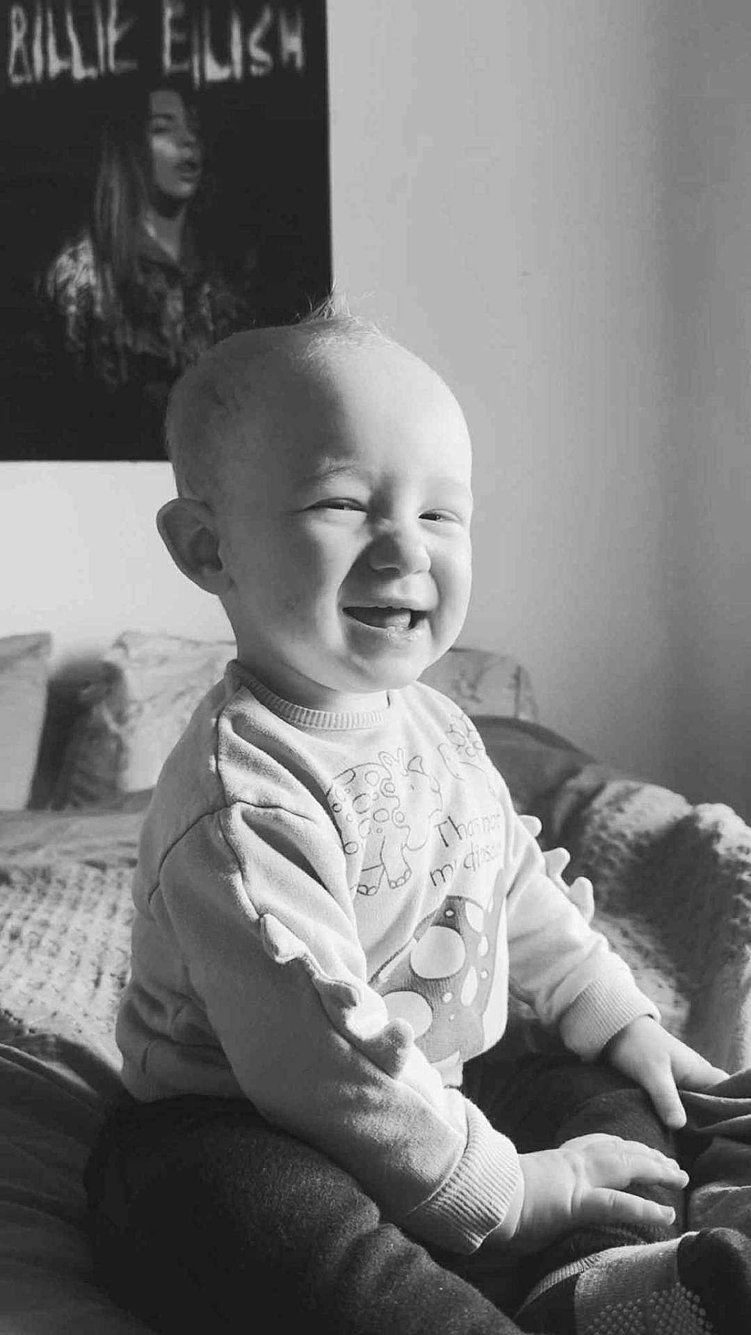 Tylan is registered to the contest to win money with this photo: baby, smiling, child, indoor, bed, clothing, sitting, portrait, black_and_white, happiness, joy, light, texture, poster, room, cozy, soft_light, expression, person, cute