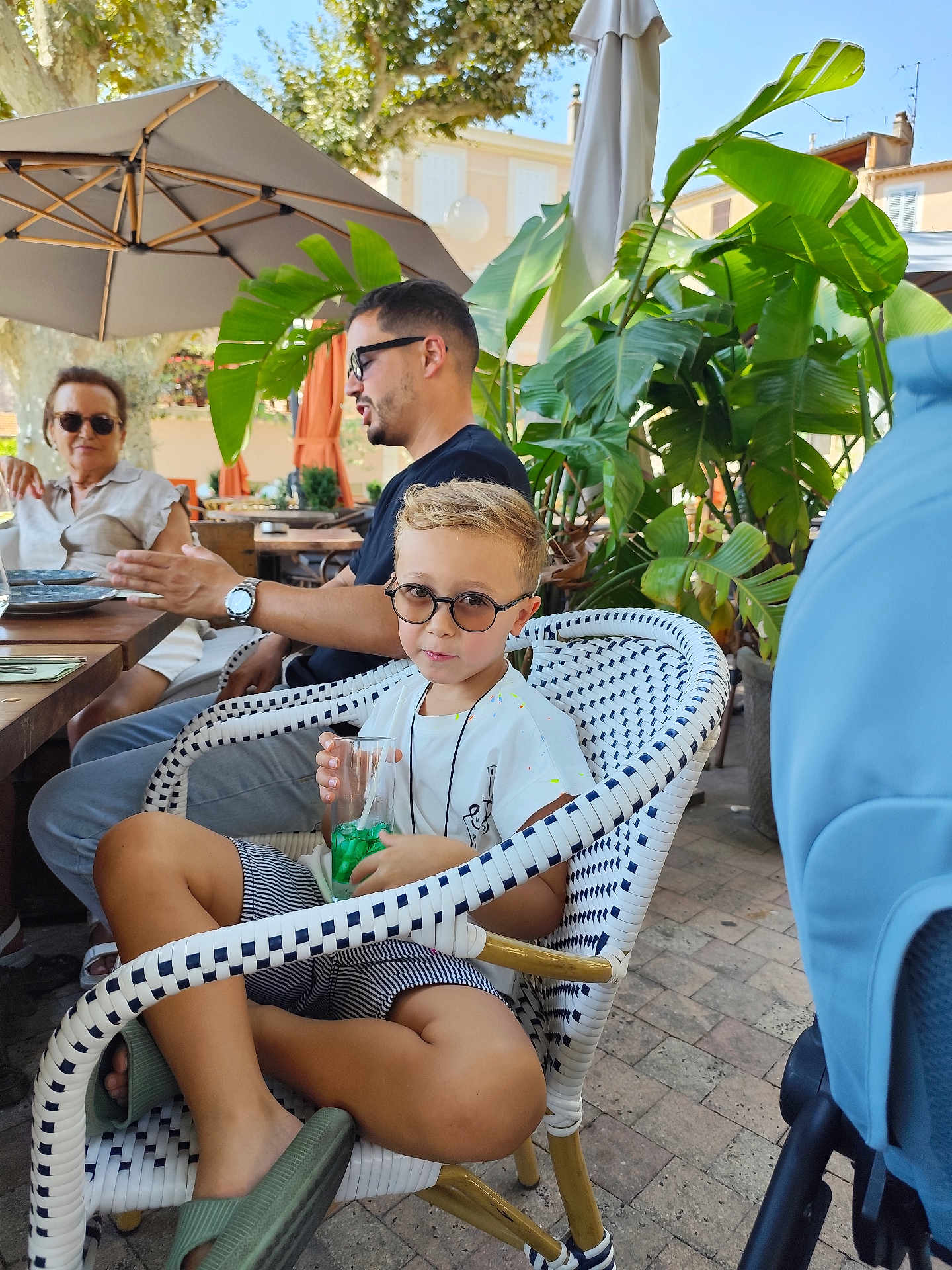 Adam participe au concours pour gagner de l'argent avec cette photo : child, boy, glasses, drink, chair, outdoor, people, umbrella, table, plants, greenery, casual, summer, relaxing, sitting, footwear, conversation, daylight, cafe, pavement