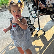 Roszalia is registered to the contest to win money with this photo: child, toddler, baby, lollipop, smiling, outdoor, sunlight, stroller, dress, striped_clothing, hair_bows, curly_hair, sparkly_shoes, standing, walking, pavement, grass, shadow, portrait, family_outing