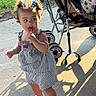 child, toddler, baby, lollipop, smiling, outdoor, sunlight, stroller, dress, striped_clothing, hair_bows, curly_hair, sparkly_shoes, standing, walking, pavement, grass, shadow, portrait, family_outing
