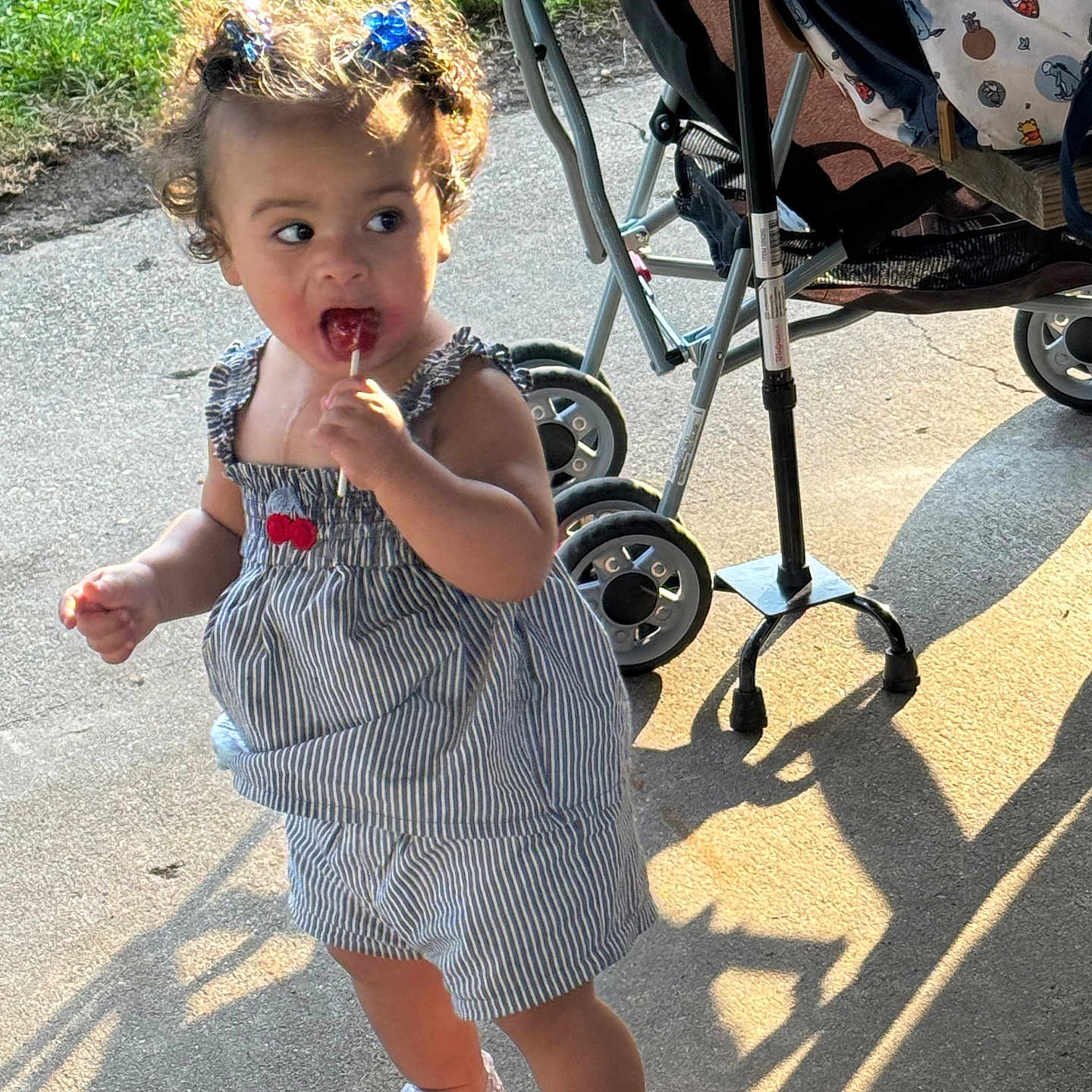 Roszalia is registered to the contest to win money with this photo: child, toddler, baby, lollipop, smiling, outdoor, sunlight, stroller, dress, striped_clothing, hair_bows, curly_hair, sparkly_shoes, standing, walking, pavement, grass, shadow, portrait, family_outing