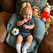 Maddox is registered to the contest to win money with this photo: baby, child, blue_cushion, plush_toys, toy_balls, soccer_ball, basketball, football, baby_clothing, white_socks, smiling, indoor, couch, brown_couch, cute, infant, happy, portrait, seated, playtime
