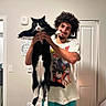 cat, black_and_white_cat, person, man, curly_hair, smiling, indoor, bathroom, door, mirror_selfie, pet, animal, holding, standing, long_cat, whiskers, paws, anime_shirt, casual_clothing, portrait