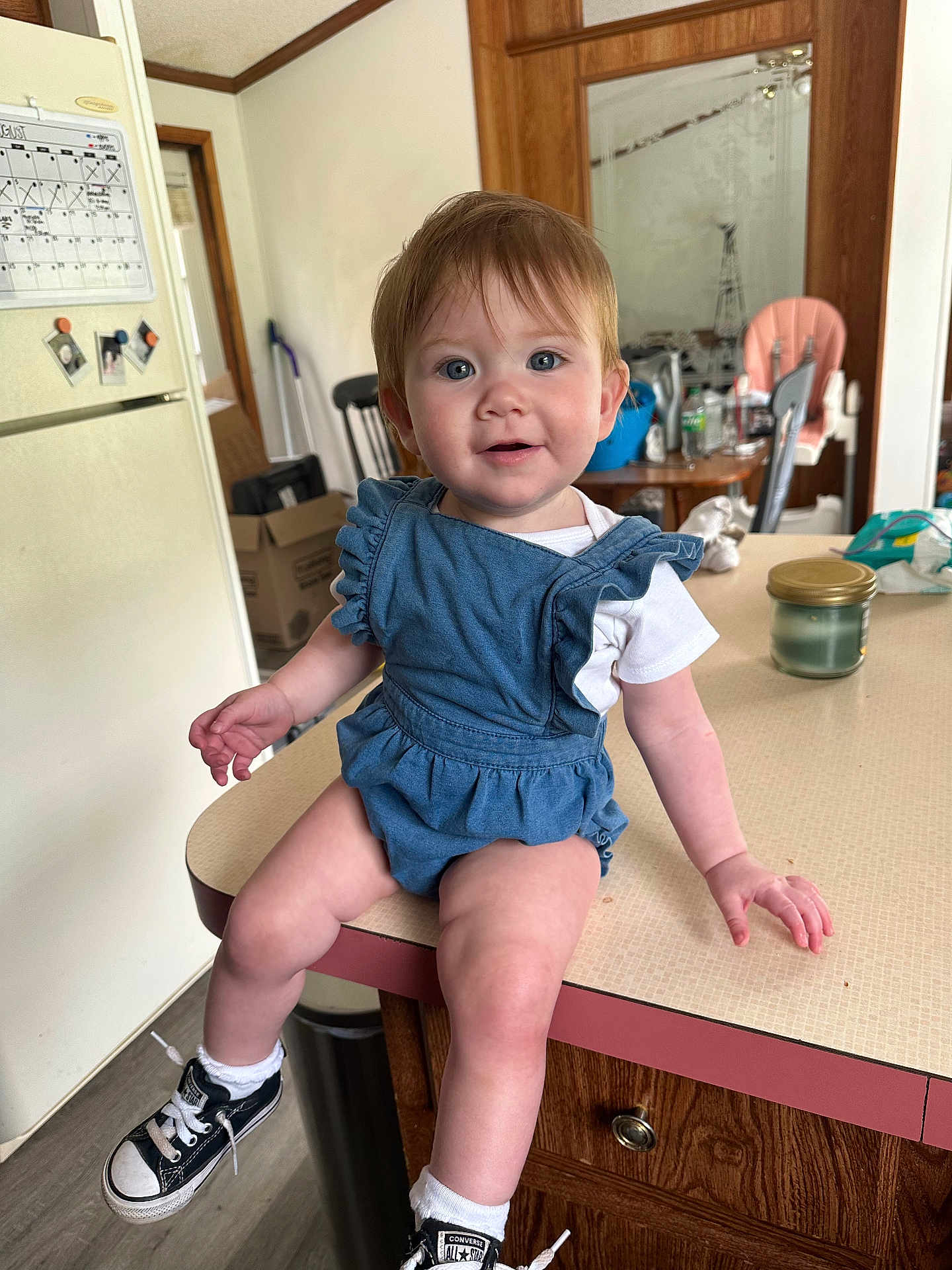 Nova is registered to the contest to win money with this photo: toddler, child, denim_romper, sneakers, converse, kitchen, counter, table, chair, refrigerator, calendar, bottle, candle, wood_cabinet, floor, indoor, smiling, face, person, clothing