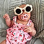 baby, sunglasses, pacifier, pink_outfit, cherry_pattern, bed, blanket, infant, cute, child, fashion, accessory, indoor, relaxing, portrait, person, headwear, sleepwear, skin, hand