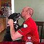 Kash is registered to the contest to win money with this photo: affection, black_dog, chair, clock, dog, embrace, furniture, home, human_animal_interaction, indoor, kitchen, love, man, pet, phone, red_shirt, table, wall, water_bottle, white_paw