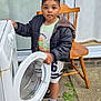 appliance, black_jacket, child, concrete, curious, curtains, door, expression, face, footwear, greenery, hand, outdoor, person, shorts, sneakers, washing_machine, window, wooden_chair, young