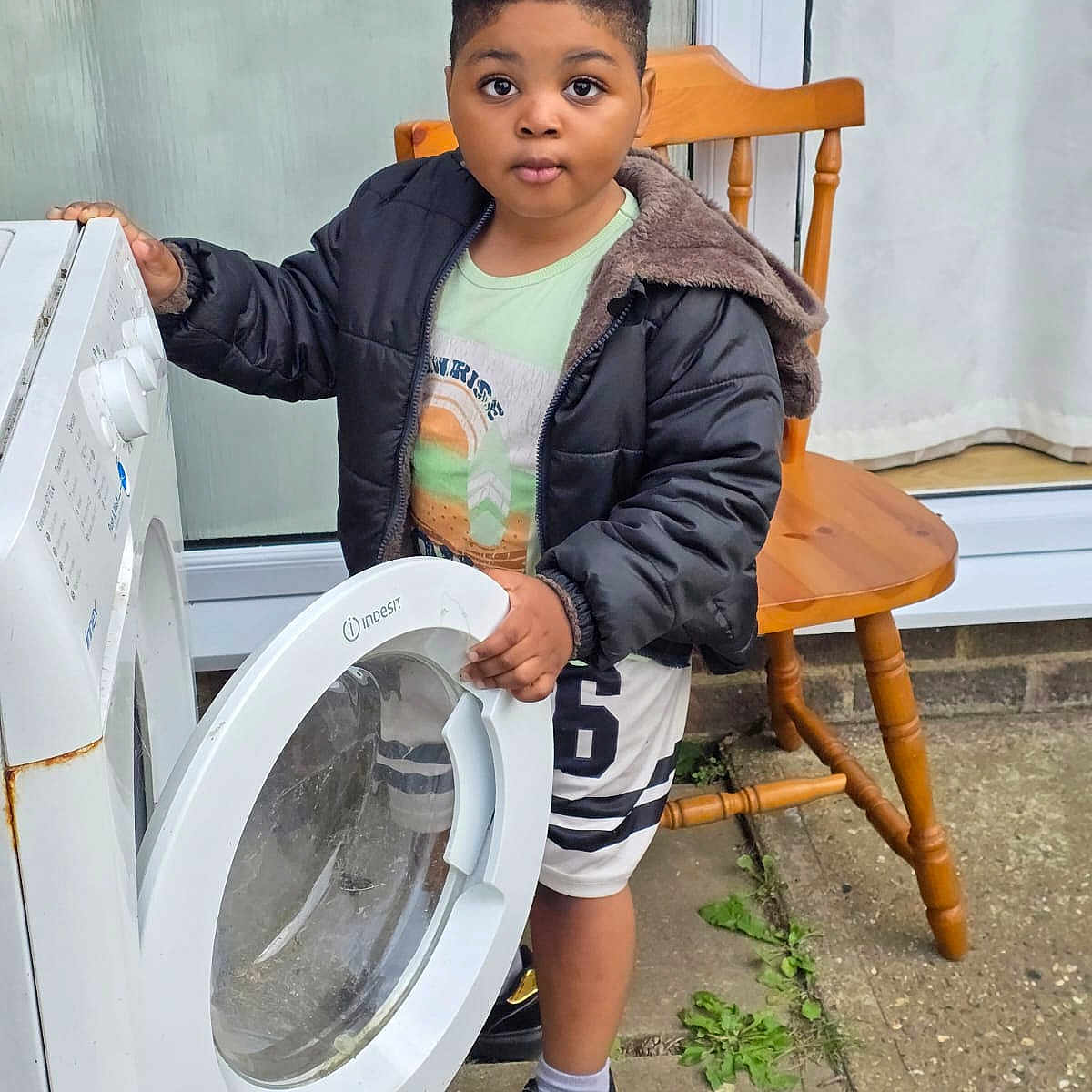 Washington is registered to the contest to win money with this photo: appliance, black_jacket, child, concrete, curious, curtains, door, expression, face, footwear, greenery, hand, outdoor, person, shorts, sneakers, washing_machine, window, wooden_chair, young