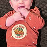 Braylee is registered to the contest to win money with this photo: baby, infant, smiling, hands, rust_colored_clothing, mushroom_pattern, patch, pizza, deep_dish_cards, lying_down, brown_background, happy, cute, chewing_fingers, face, person, child, closeup, indoors, portrait