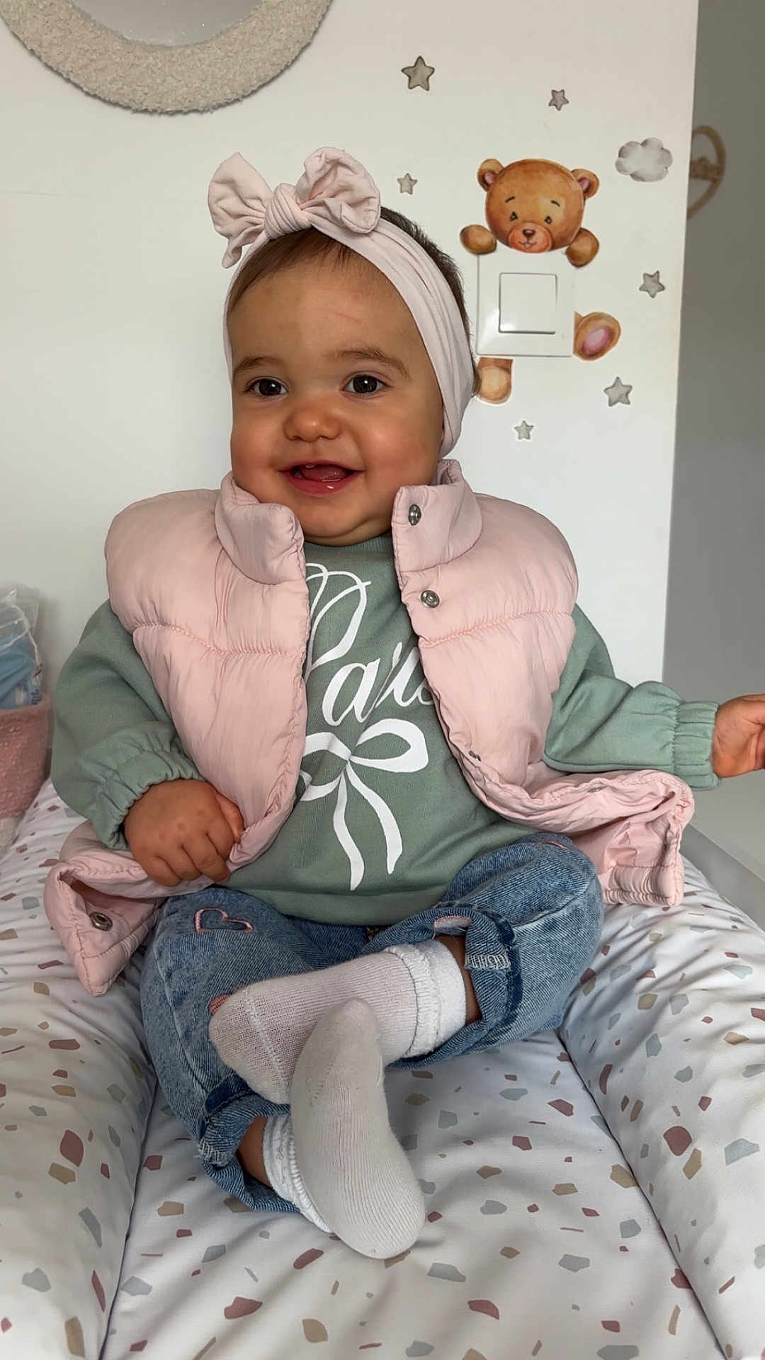 Victoire participe au concours pour gagner de l'argent avec cette photo : baby, child, smiling, headband, pink_vest, green_sweater, denim_jeans, white_socks, cushion, patterned_bedding, teddy_bear_wall_decor, stars_wall_decor, indoor, cute, happy, portrait, seated, infant, head, clothing