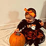 baby, child, toddler, pumpkin, halloween, orange_bow, black_dress, smiling, indoor, blanket, wall, floor, cute, festive, holiday, sitting, person, happy, headband, celebration