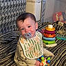 baby, pacifier, bib, toy, colorful_toys, striped_blanket, smiling, sitting, indoor, child, cute, playtime, face, infant, happy, head_tilt, clothing, soft_toy, background, home