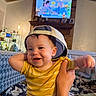 baby, child, smiling, yellow_onesie, cap, backwards_hat, hand, living_room, couch, television, brick_fireplace, person, indoor, happy, playful, portrait, infant, cute, family, casual