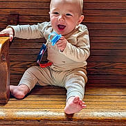 Skyler is registered to the contest to win money with this photo: baby, child, pacifier, wooden_bench, smiling, happy, indoor, pajamas, barefoot, infant, cute, seated, wood_texture, light_skin, short_hair, playful, one_person, holding_object, casual, relaxed