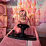 child, baby, toddler, pink, princess, castle_backdrop, toy_car, steering_wheel, pink_dress, headband, bow, birthday_party, balloon, decorations, stacked_blocks, glitter, portrait, indoor, celebration, smiling