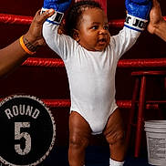 Kyrie is registered to the contest to win money with this photo: baby, boxing_gloves, boxing_ring, child, white_onesie, socks, adult_hands, round_5_sign, blue_gloves, red_background, stool, bucket, sport, indoor, cute, determined, standing, person, portrait, funny