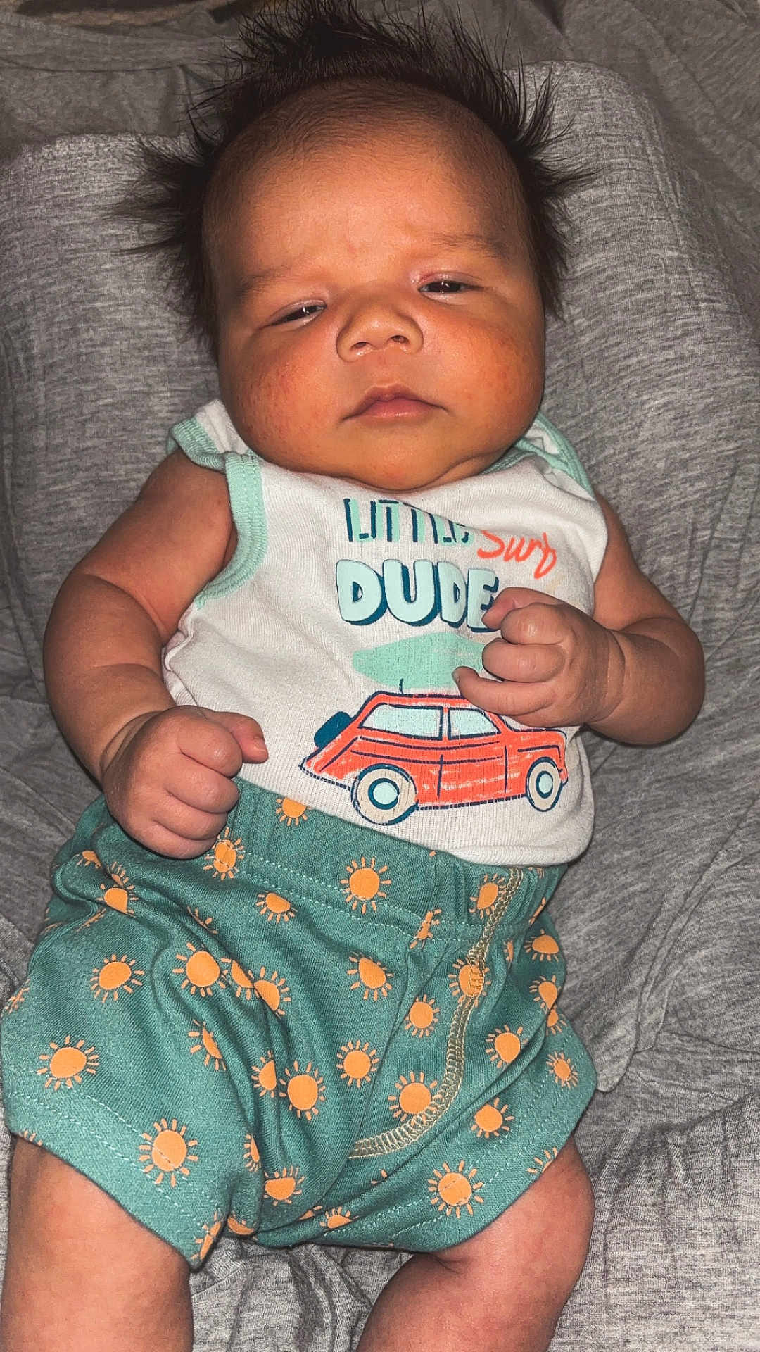 Knox is registered to the contest to win money with this photo: baby, infant, child, face, hair, clothing, tank_top, shorts, pattern, sun_pattern, car_print, fist, expression, laying, blanket, soft, skin, cute, indoors, portrait