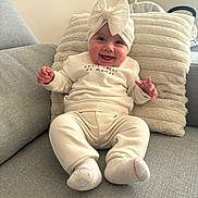Lina participe au concours pour gagner de l'argent avec cette photo : baby, smiling, headband, bow, white_clothing, socks, couch, pillow, indoor, child, happy, cute, infant, person, seated, soft_texture, cozy, portrait, face, hands
