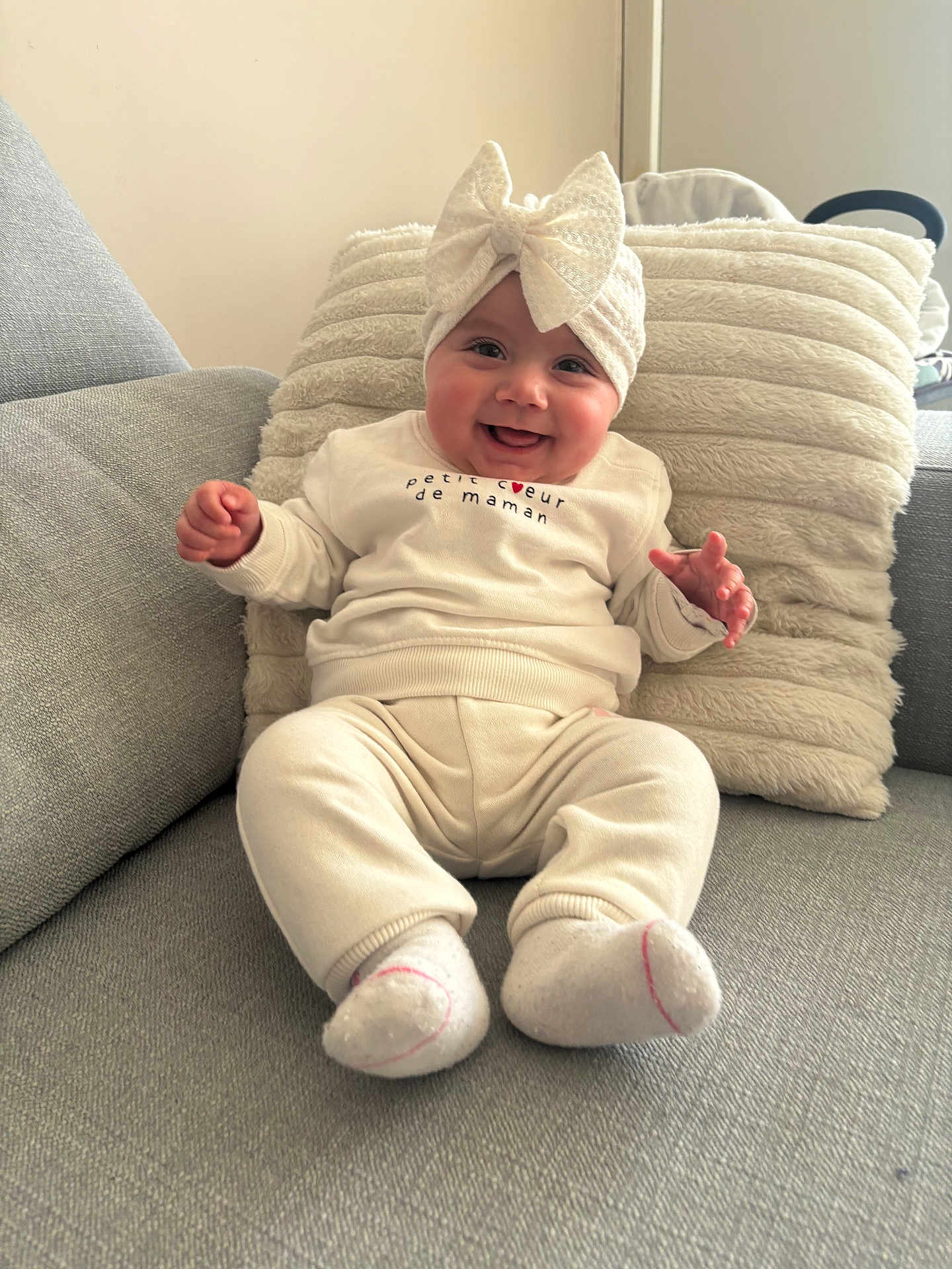 Lina participe au concours pour gagner de l'argent avec cette photo : baby, smiling, headband, bow, white_clothing, socks, couch, pillow, indoor, child, happy, cute, infant, person, seated, soft_texture, cozy, portrait, face, hands