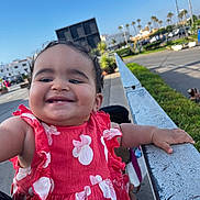 Raneem participe au concours pour gagner de l'argent avec cette photo : toddler, child, smiling, red_dress, outdoor, daylight, urban, railing, hand, person, happy, curly_hair, baby, holding, street, sky, car, plants, sidewalk, parked_cars