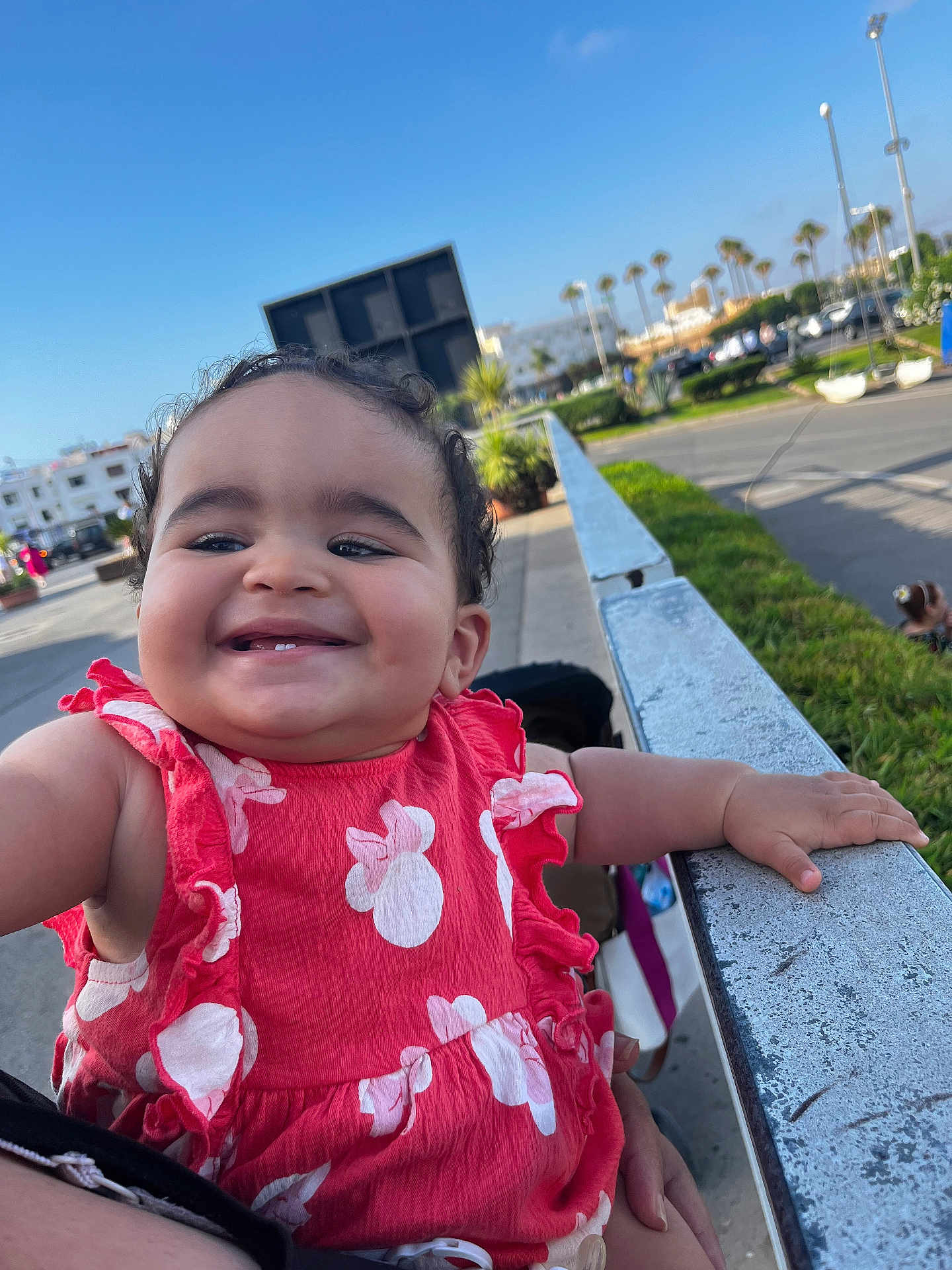 Raneem participe au concours pour gagner de l'argent avec cette photo : toddler, child, smiling, red_dress, outdoor, daylight, urban, railing, hand, person, happy, curly_hair, baby, holding, street, sky, car, plants, sidewalk, parked_cars