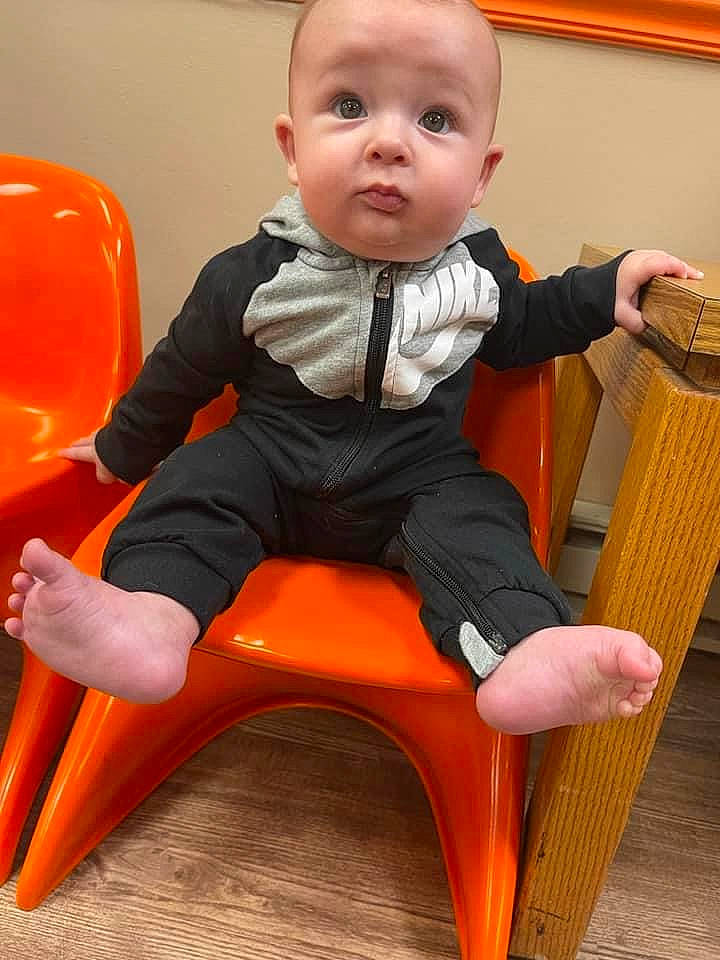 Nolan is registered to the contest to win money with this photo: arm, baby, baby_toddler_clothing, chair, child, comfort, face, finger, foot, head, knee, lap, leg, orange, person, sitting, sleeve, surprise, t_shirt, thigh