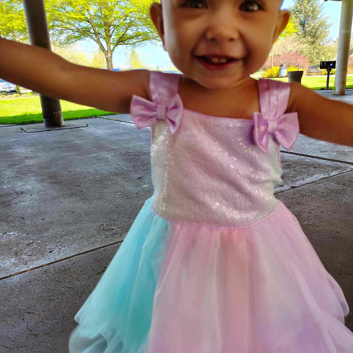 Nova is registered to the contest to win money with this photo: bows, child, closeup, concrete_floor, cute, daytime, dress, greenery, happy, outdoor, pastel_colors, pavilion, person, playful, portrait, smiling, sunlight, toddler, trees, young_child