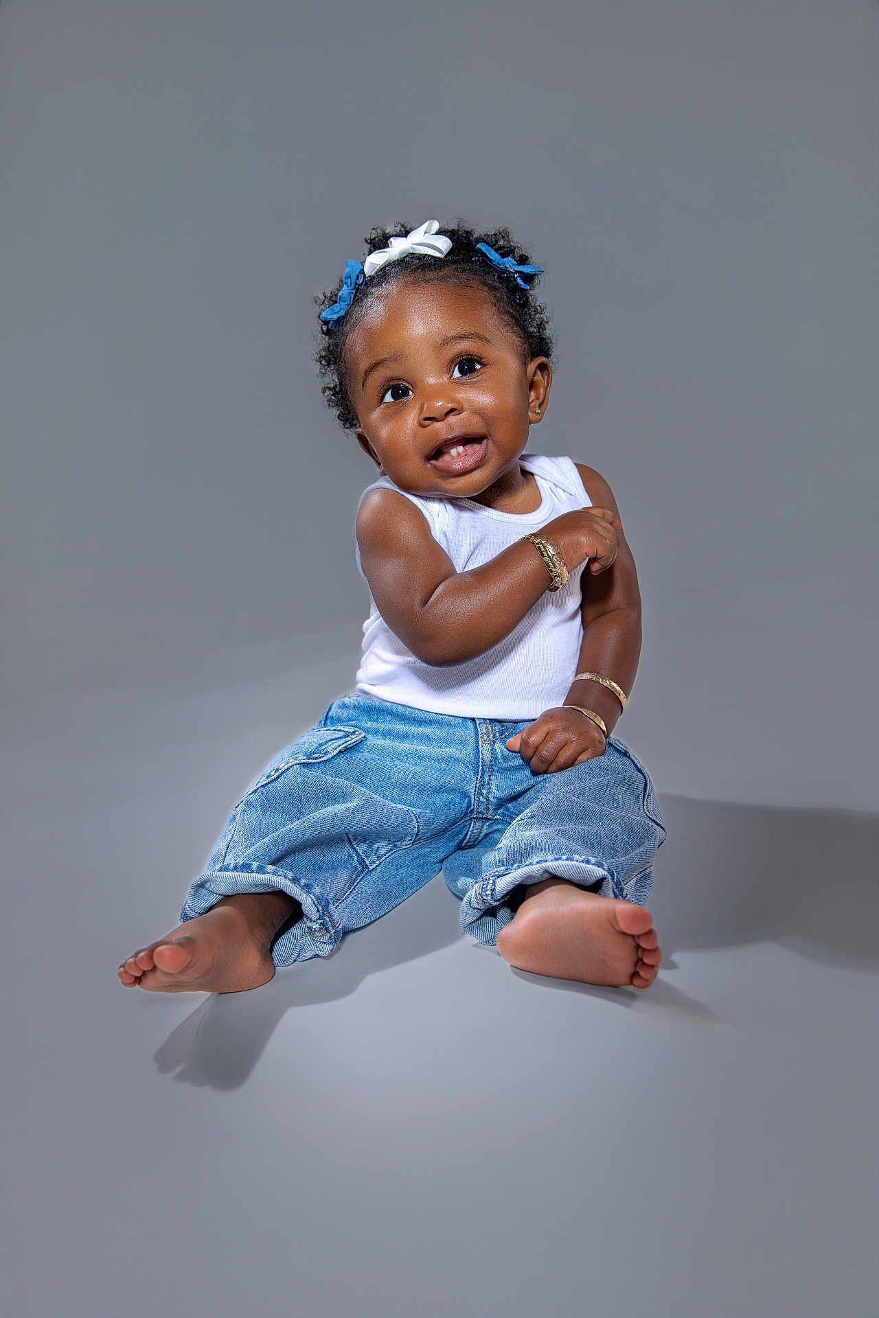 Leylani is registered to the contest to win money with this photo: baby, child, smiling, jeans, tank_top, barefoot, bracelets, hair_bows, curly_hair, studio, portrait, sitting, cute, happy, expression, skin, foot, fashion, accessory, one_person